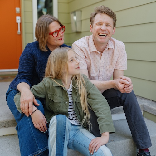 Family sitting on front porch steps of green home with orange door.