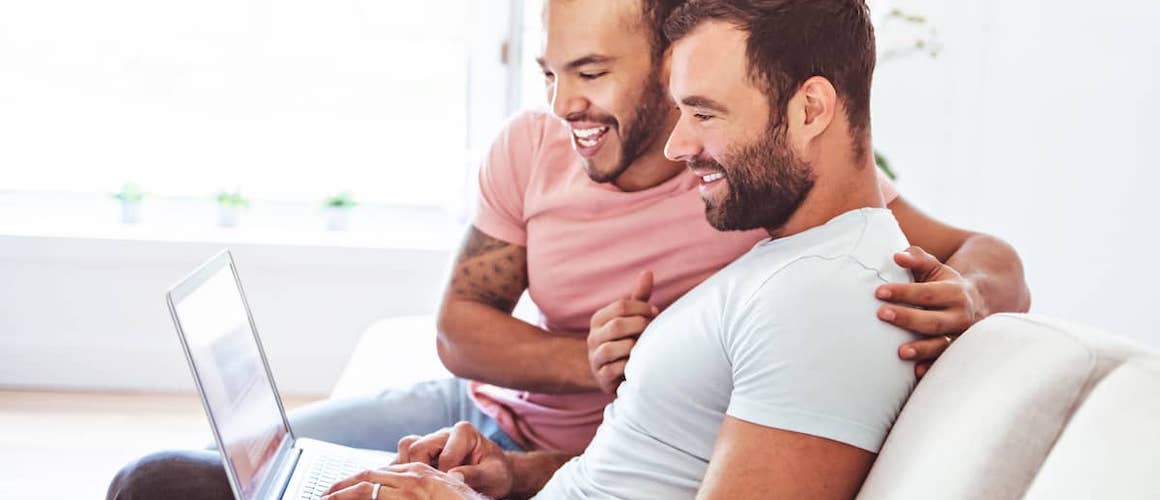 Young male couple smiling and looking at a laptop together while sitting on the couch.