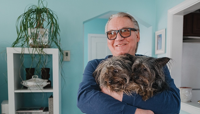 Man wearing orange glasses holding two small dogs in light blue room.