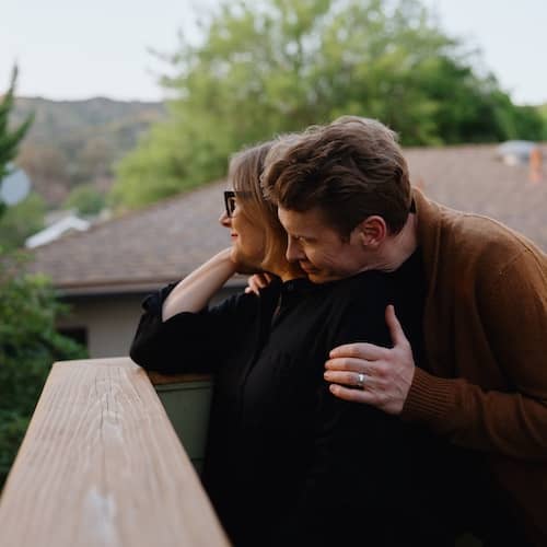 Couple holding each other while they look off wooden deck into the woods.