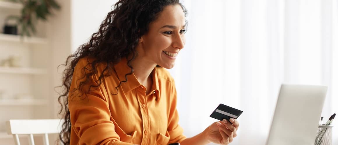 Young woman smiling widely at her laptop while holding a credit card.