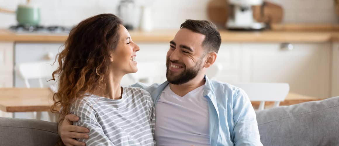 A couple smiling and talking while sitting on the couch in their house.