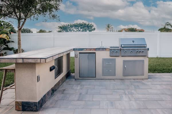 Outdoor kitchen on gray stone flooring in a backyard including a grill.