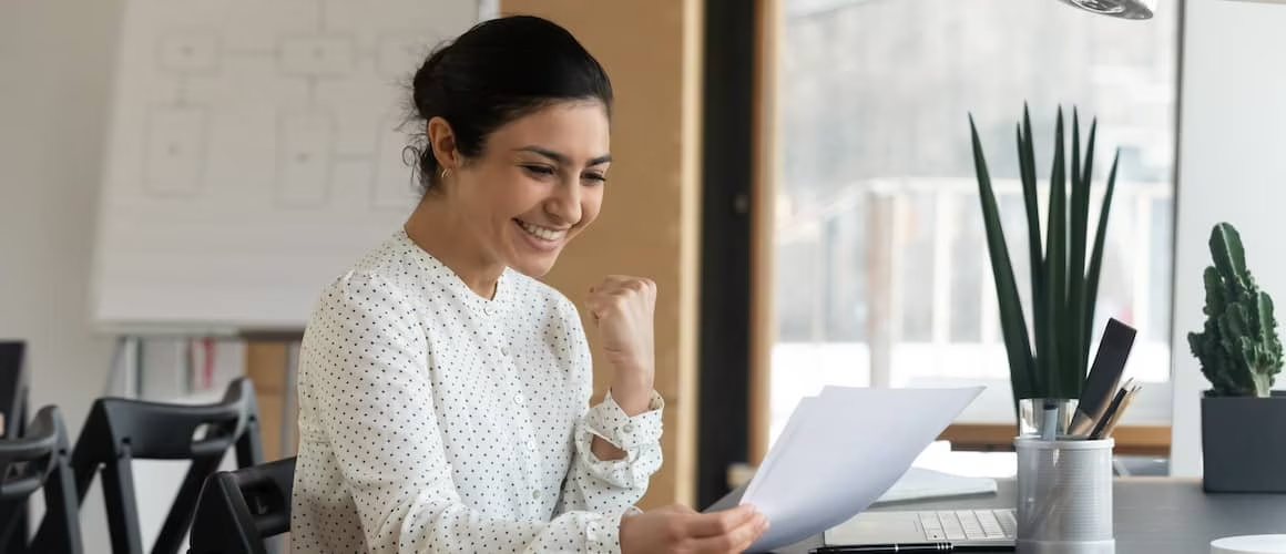 Woman in a white and polka dot shirt looking excited and happy while holding a sheet of paper.