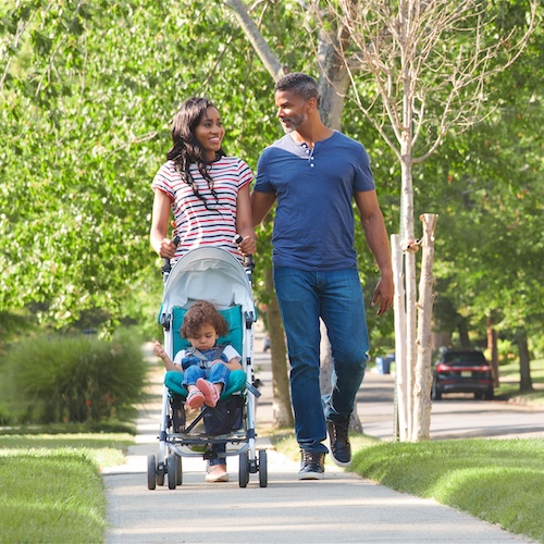 Couple Push Daughter In Stroller As They Walk Along Suburban Street.