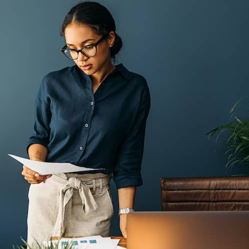 A woman wearing glasses stands at a desk, working on a laptop computer and looking on a piece of paper.