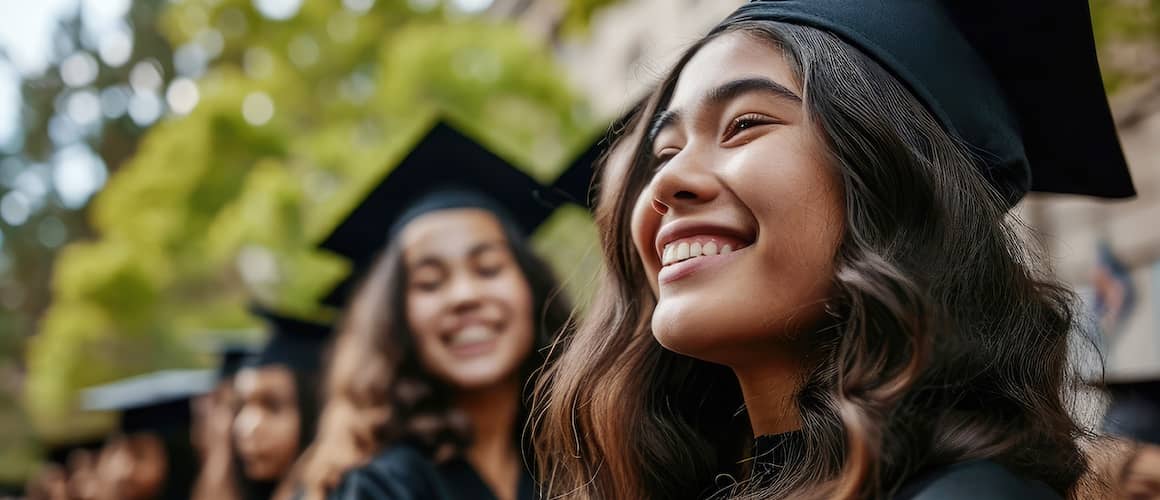 Smiling young woman college graduate in black graduation gown and cap surrounded by other graduates.