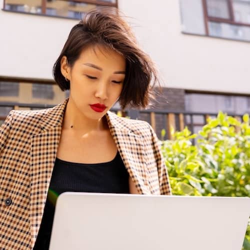 Woman working on laptop outside of apartment building, wearing a blazer loosely on shoulders.