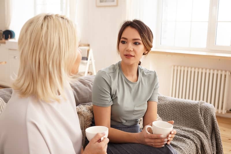 Woman talking to her daughter while they both drink tea or coffee on the couch.