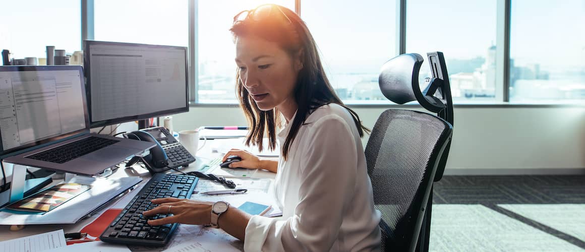 Professional woman typing something with multiple screens in front of her.