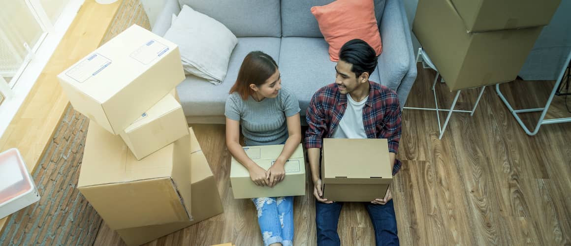 A young Asian couple smiles as they unpack cardboard boxes in a warmly lit living room, creating a cozy scene of settling into their new home.