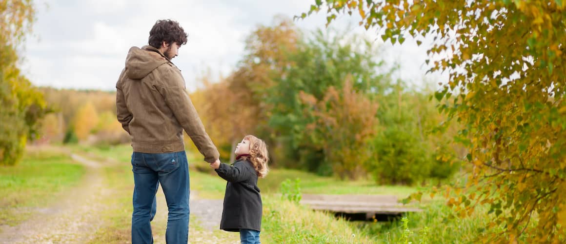 Smiling father in tie affectionately walking down tree-lined street holding hands with his young daughter.