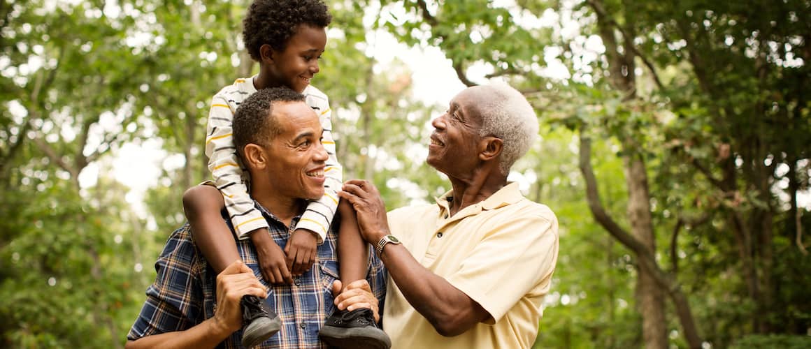 Multigenerational African American family of three standing in a wooded area and smiling.