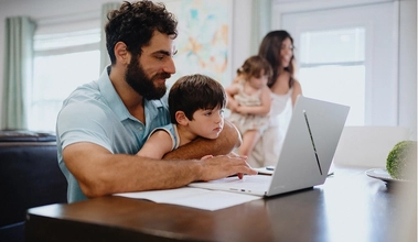 A man sitting at a table on his laptop researching amortization schedule while his son sits in his lap.