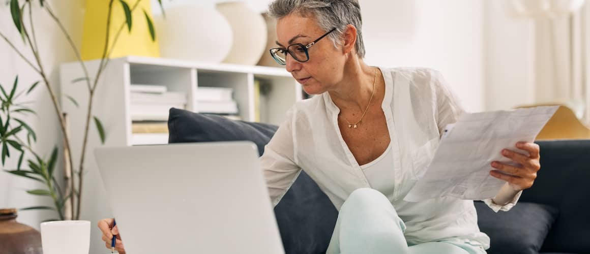 Woman with silver haired pixie cut working on finances with open laptop on couch.