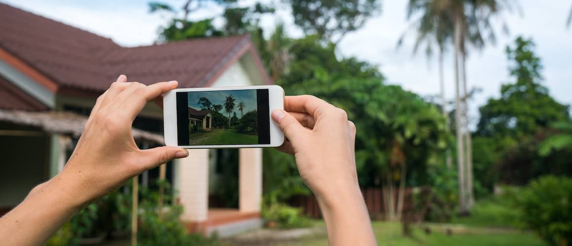 Hands holding a smartphone photographing a house, possibly related to real estate or property viewing.