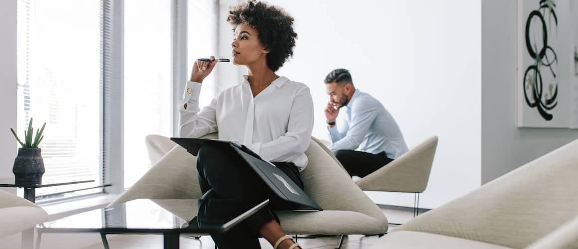 A woman sitting in a fancy room thinking something with a pen in hand while a man in background is talking on phone.