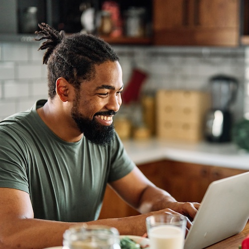 Man smiling while working on laptop in kitchen while eating breakfast.