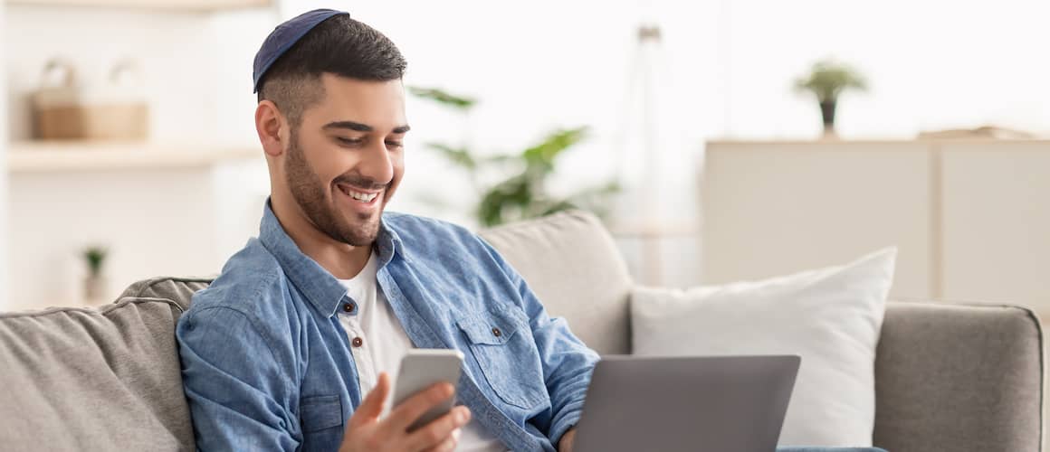 Young man wearing a yarmulk holds his phone up in one hand and smiles at his laptop on his lap.