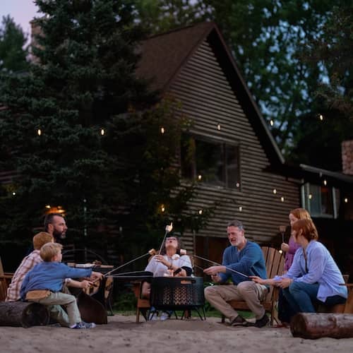 A family roasting marshmallows over a metal fire pit behind their home.