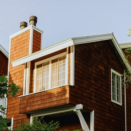 The upper portion of a two-story house with brown shingle siding, white trim, and a large window beneath a sloped roof, with twin chimney stacks rising above against a clear sky.