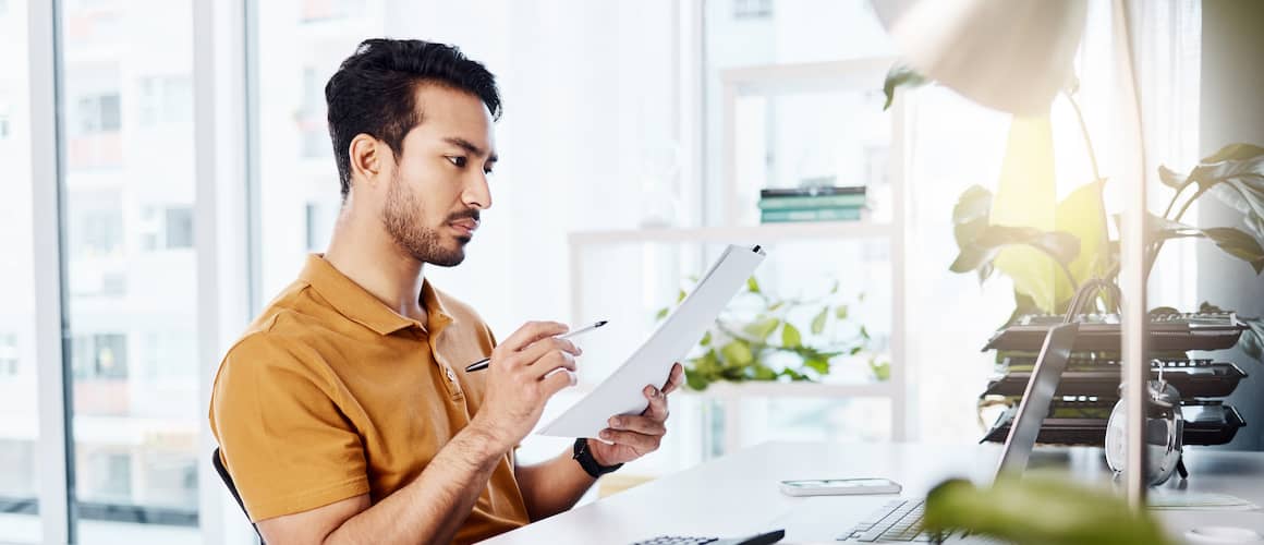 Man in yellow shirt studying paperwork and calculating budget in bright, sunlight office with plants.