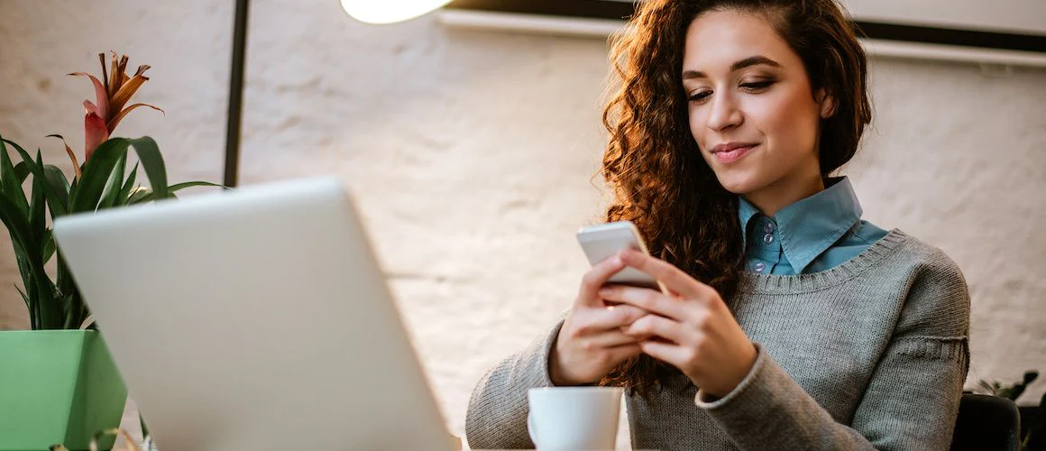 Woman in front of open laptop, looking at mobile phone.