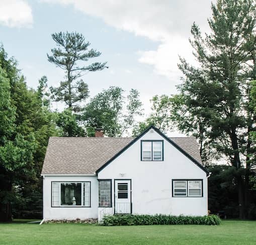 Small white home surrounded by tall trees on a large lawn.