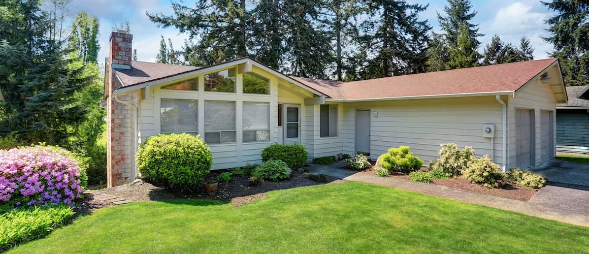 White, one story house with brick chimney, big window, garage and well manicured lawn.