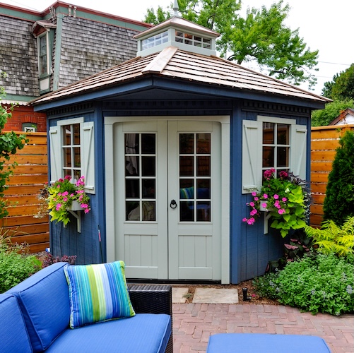 Upscale shed in backyard with double doors and windows with shutters, possibly being used as office space.