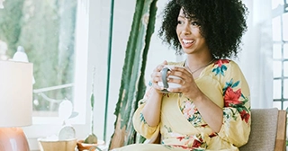 Una mujer negra sentada en una silla en su casa y bebiendo una taza de café.