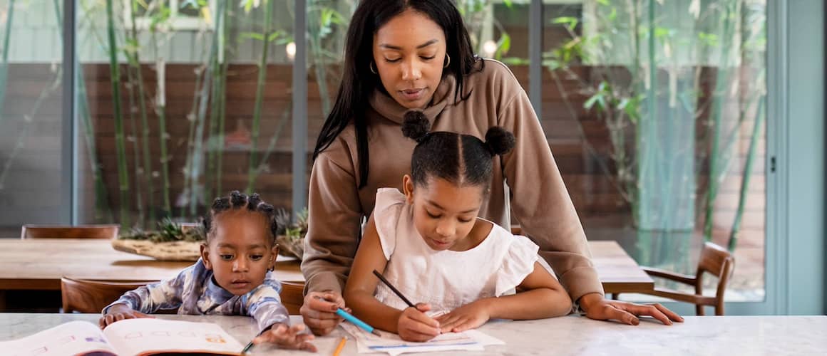 Mother helping young daughters with school work at large table in open room with patio in background.