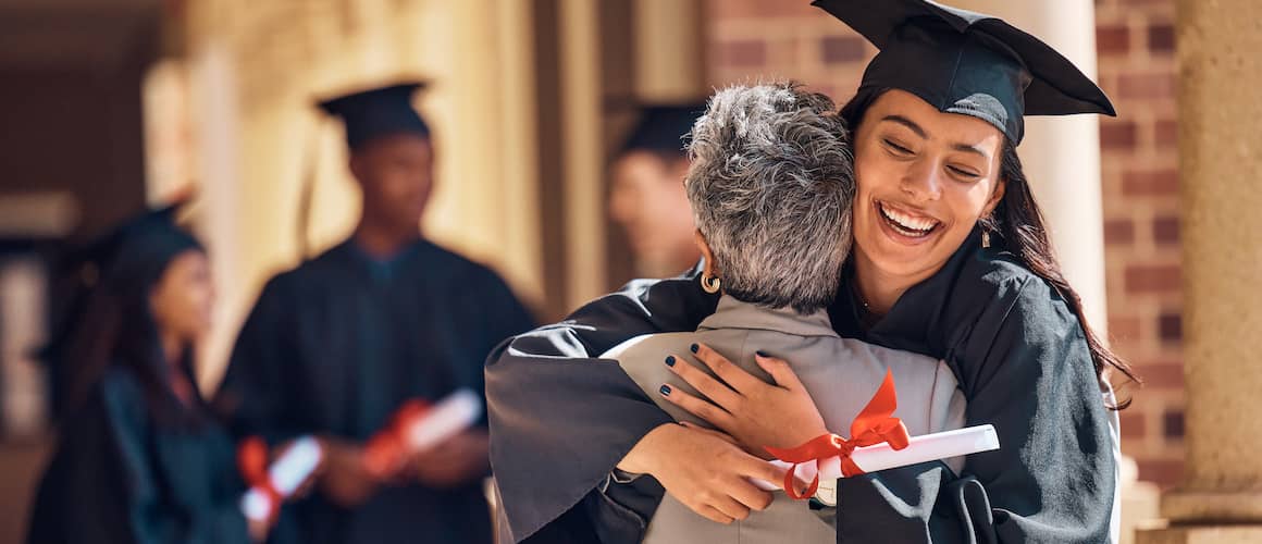 Young woman with graduation cap and gown hugging older woman and smiling.