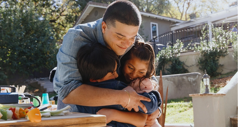 A man with short dark hair smiles and leans over to hug two kids while standing outside in the yard.