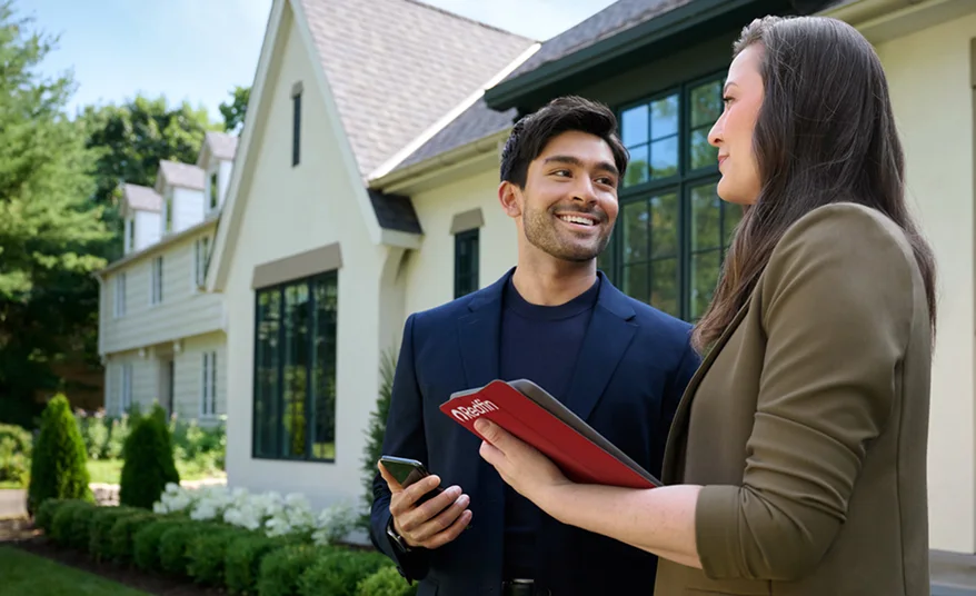 Two professionals stand outside a large suburban home with light siding and tall windows. The man smiles while holding a phone, and the woman holds a red Rocket-branded folder as they talk. Green shrubs and trees surround the house in the background.