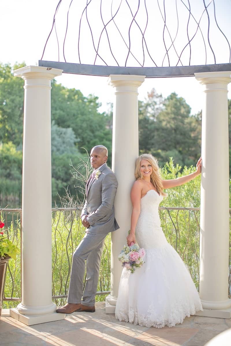 Man and woman in wedding clothes standing next to columns in front of lush greenery.