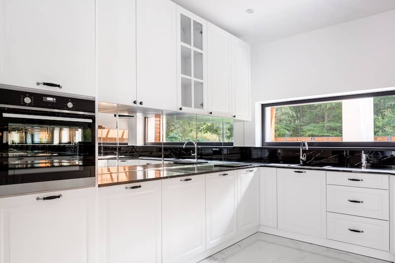Modern white kitchen with mirrored backsplash reflecting picture window looking outside to lush green yard.