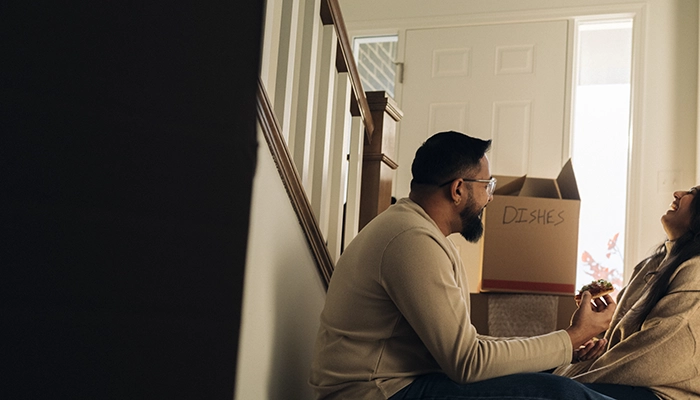 Couple holding hands laughing sitting next to pizza box on hallway floor.