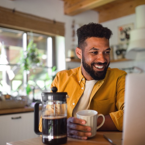 Man smiling, drinking coffee and looking at open laptop in kitchen.