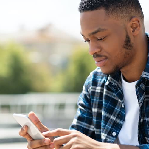 A black man in a blue plaid shirt looking at his phone on a sunny day.