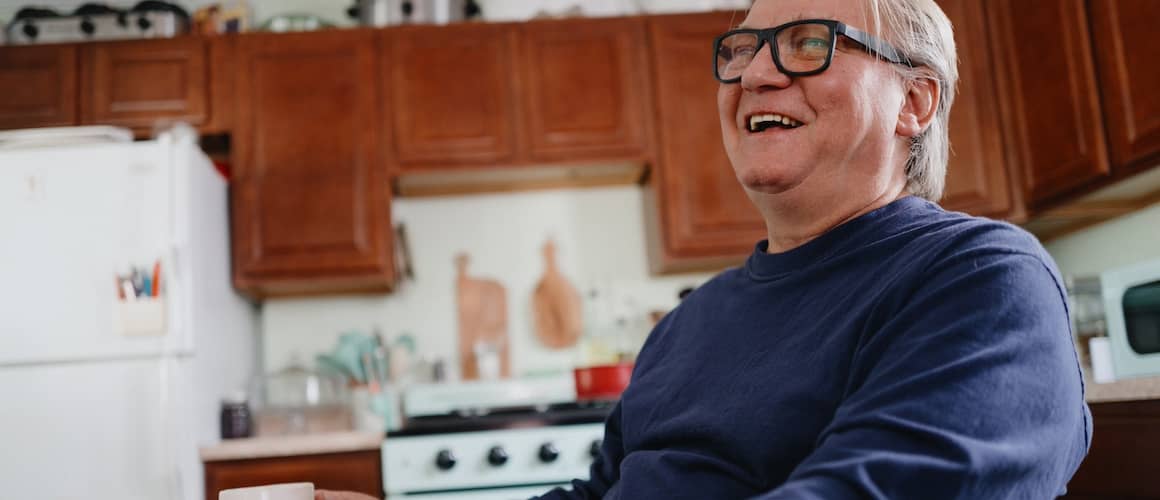 Older man sitting in kitchen smiling.