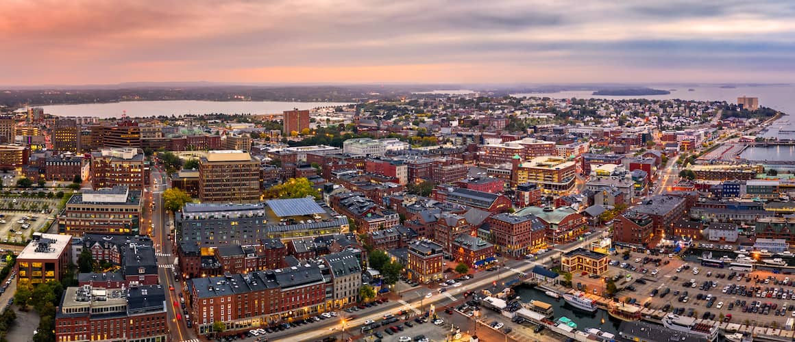 Portland Maine at dusk lit up by building and streetlights and with water in the background.