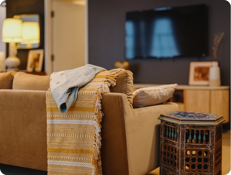 Living room with a tan sofa, patterned blanket, colorful side table, and soft lighting.