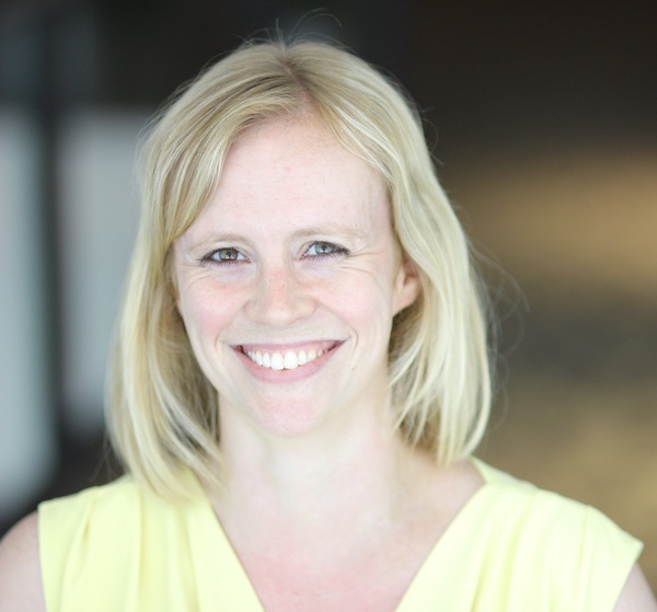 Blonde woman smiling in yellow shirt with a blurred background.