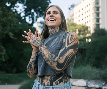  A woman with tattoos talking outside surrounded by trees and a city view in a park.