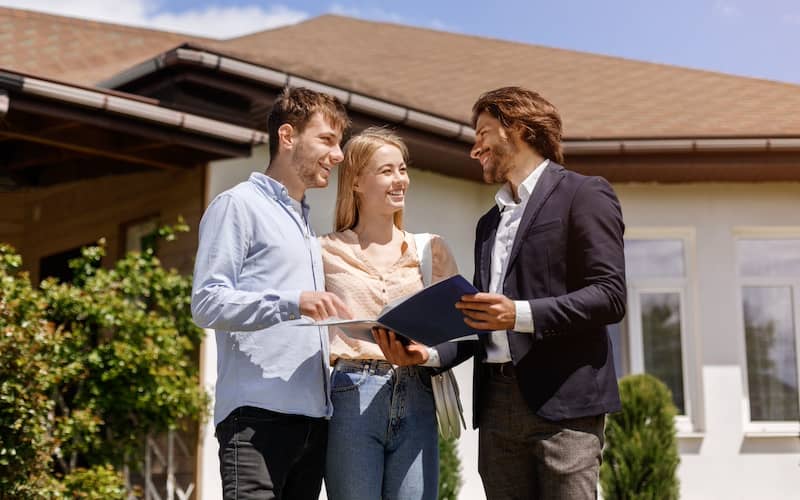 Real estate agent standing with couple in front of a house looking at documents in a folder.