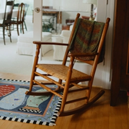 A wooden rocking chair with a woven seat and patterned cushion sits on a rug in a bright living space, with a dining area visible in the background.