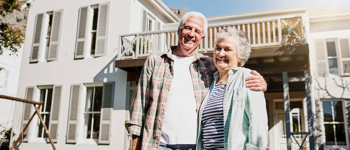 An elderly couple smiling outside their house indicating a peaceful living.