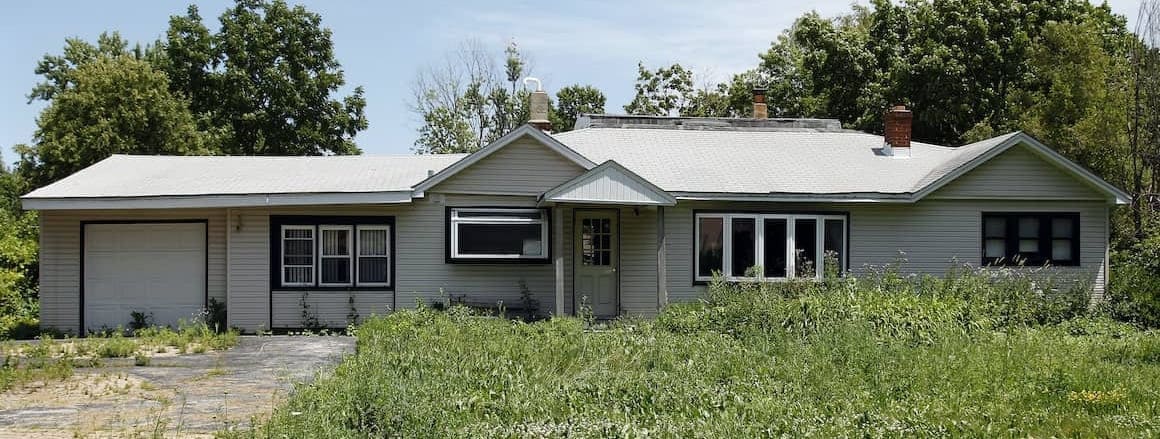 Exterior view of distressed grey ranch-style family house with an unkempt overgrown lawn in disrepair.