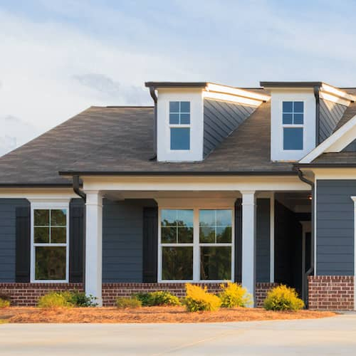 An image displaying a grey, single-story ranch-style model house with a well-maintained lawn and driveway.